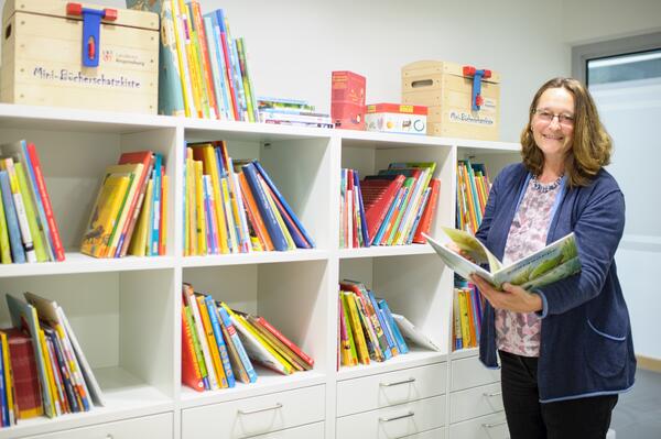 Bild vergr&ouml;&szlig;ern: Frau steht in der Bücherei und hält ein aufgeschlagenes Buch in den Händen.
