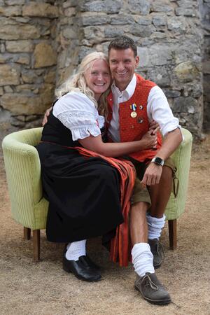 Bild vergr&ouml;&szlig;ern: Ein Paar in Tracht, das sich umarmt, sitzt auf einem gelben Sofa.