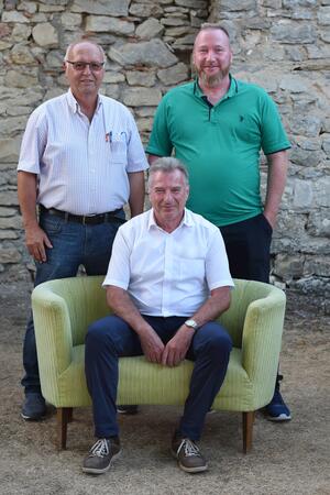 Bild vergr&ouml;&szlig;ern: Ein Mann sitzt auf einem gelben Sofa, zwei Männer stehen dahinter.