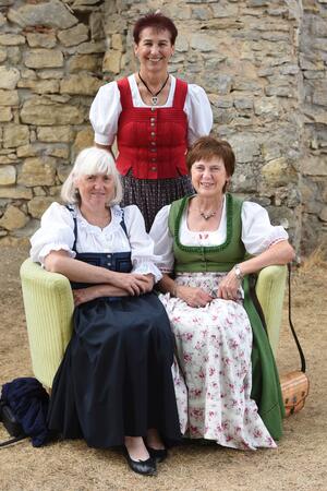 Bild vergr&ouml;&szlig;ern: Zwei Frauen in Tracht sitzen auf einem gelben Sofa und eine Frau steht dahinter.